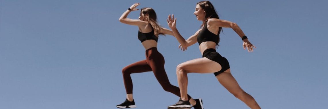 Two women in active wear leap energetically on stairs against a clear blue sky.
